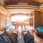 Pov view of happy couple inside minivan at sunset - Young people having fun on summer vacation - Travel,love and nature concept - Focus on feet - Warm contrast filter